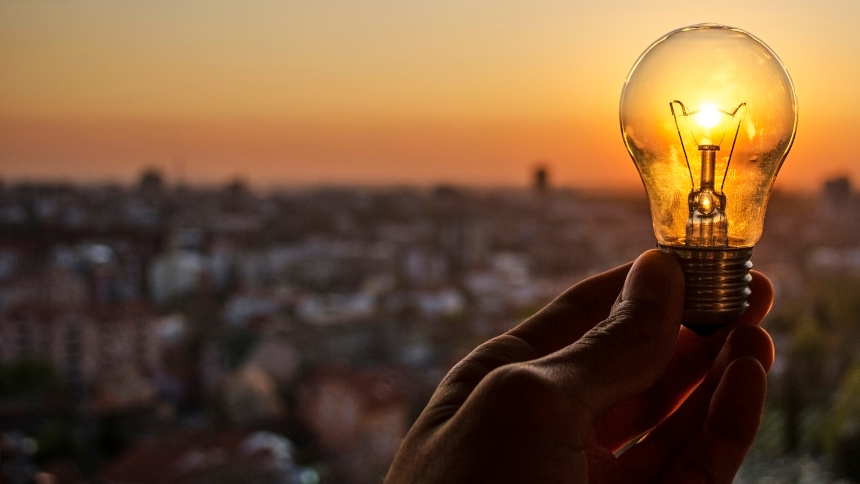 Hand holding a lightbulb with a city skyscape in the background.